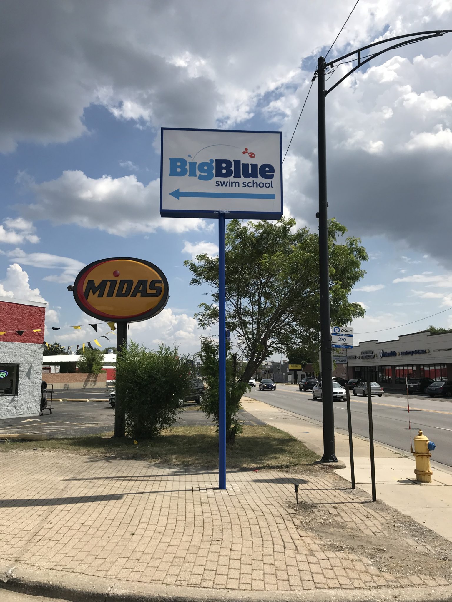 A busy intersection with business, there are two big signs. One yellow that says "Midas" and a taller white one with blue letters that says " Big Blue", representing how one can benefit from calling the best Merrillville sign company.