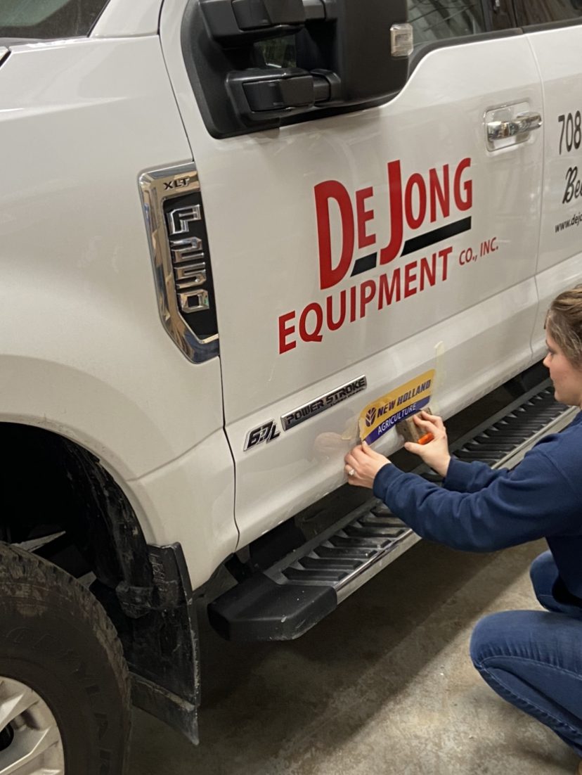 A women adding a bumper sticker to a white truck with a logo printed on, representing how one can benefit from calling the best sign company in Joliet.